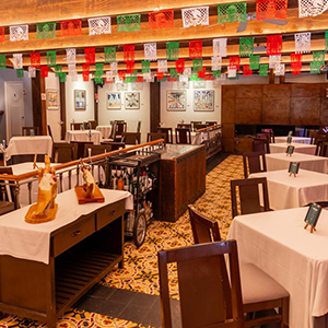 Interior de un restaurante con mesas blancas y decoración colorida, incluyendo banderitas de papel de colores colgando del techo.