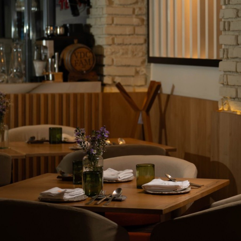 Interior de un restaurante con mesas de madera, decoradas con jarrones de flores y cristales verdes. La iluminación es suave y el ambiente es acogedor.