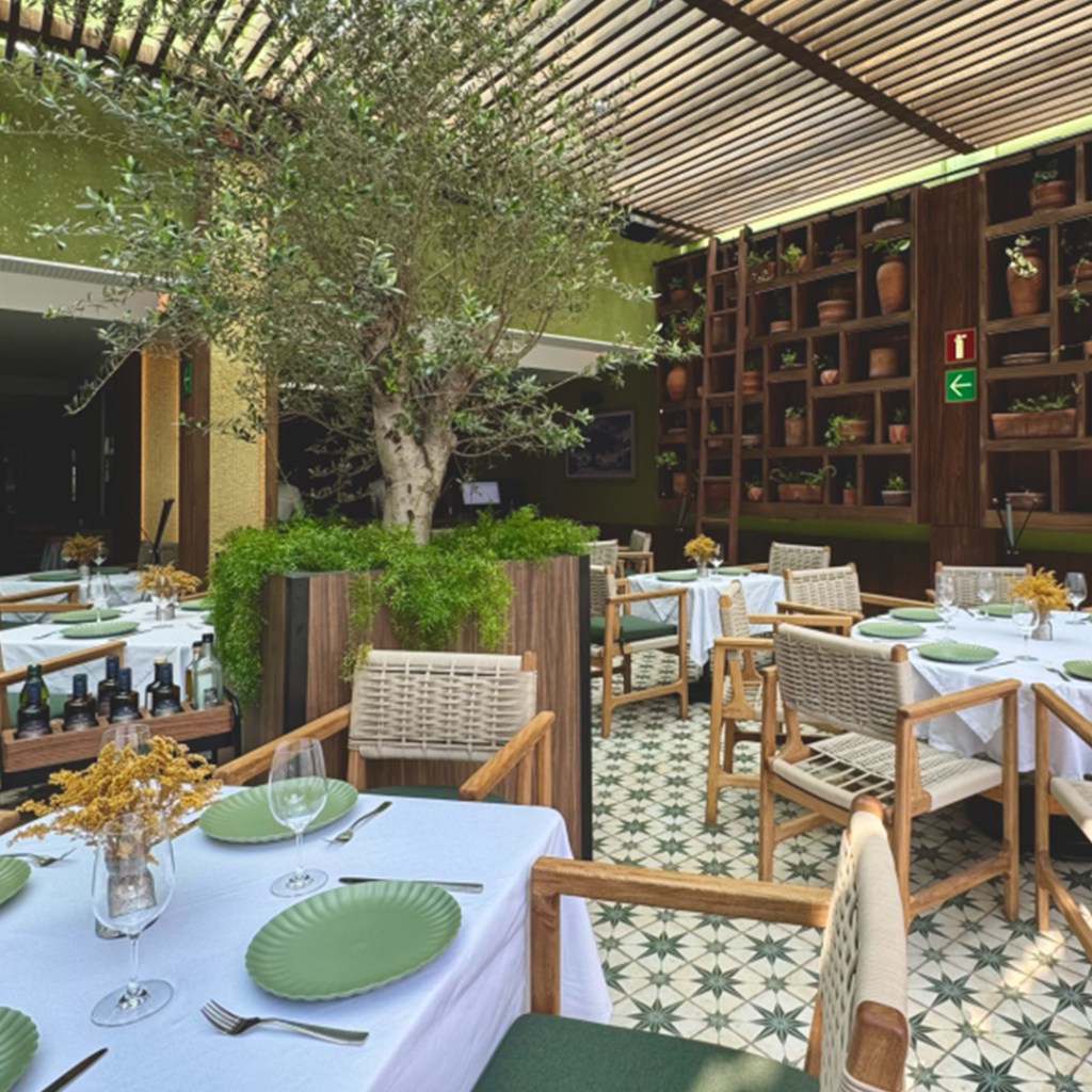 Interior de un restaurante con mesas blancas y platos verdes, decorado con plantas y un árbol en el centro, paredes de color verde y estanterías con macetas.