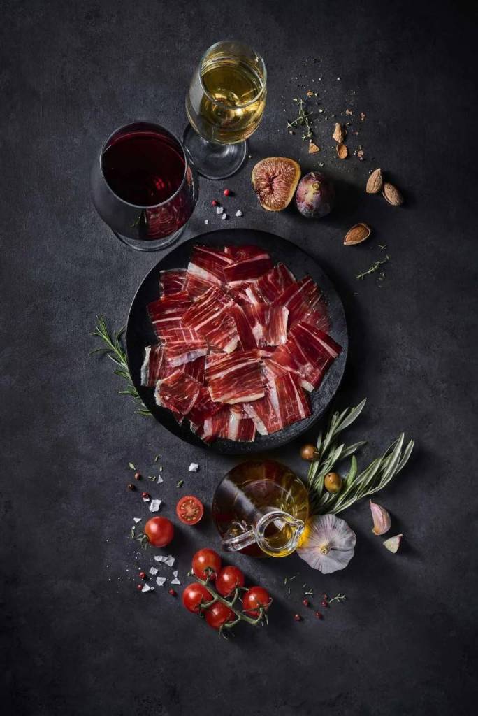 Plato con lonchas de jamón, rodeado de tomates cherry, higos, almendras, hierbas y dos copas de vino sobre fondo oscuro.