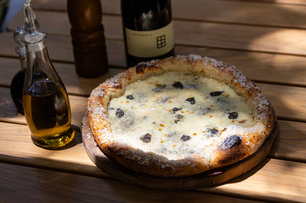 Pizza artesanal con una corteza dorada, cubierta de queso y aceitunas negras, acompañada de aceite de oliva y un vino tinto, sobre una mesa de madera.