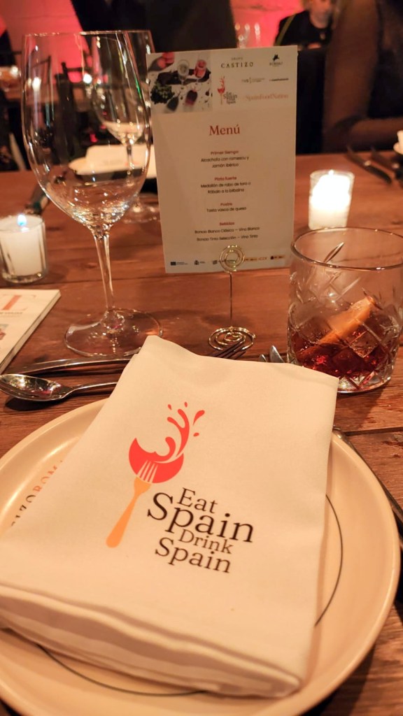 Mesa decorada para una cena, con un menú en español, una servilleta con el logo 'Eat Spain Drink Spain', un vaso con bebida y copas de vino.