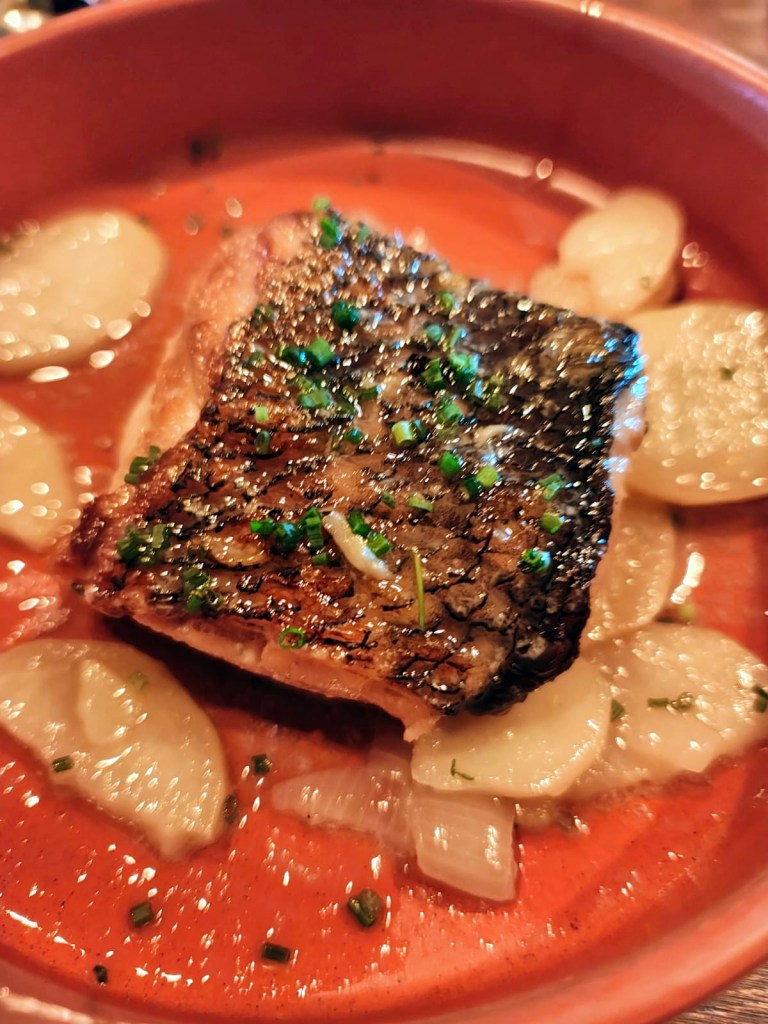 Filete de pescado con piel crujiente, servido en un caldo rojizo con rodajas de papa y cebolla, decorado con cebollino picado.