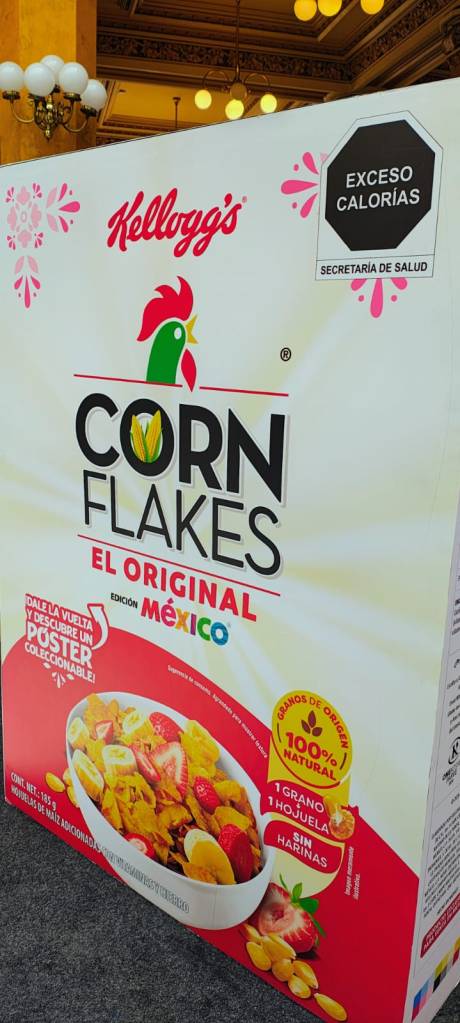 Caja de cereales Kellogg's Corn Flakes El Original, edición México, destacando la advertencia de exceso de calorías y ofreciendo una colección de postales.