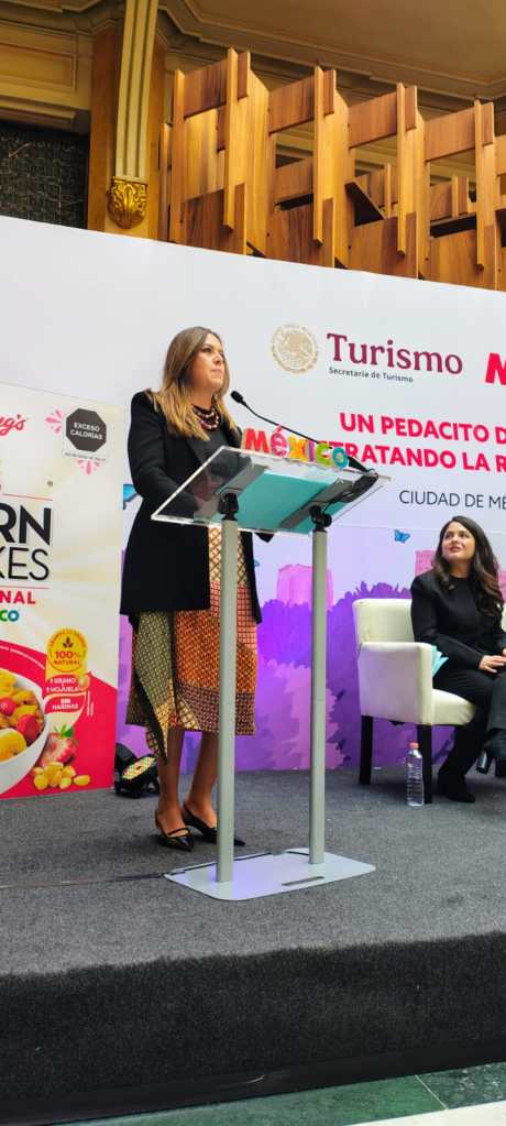 Una mujer en el escenario, hablando en un evento sobre turismo en México, con un cartel de fondo que presenta información sobre un programa de educación alimentaria.