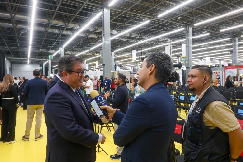 Una entrevista en un evento, con un hombre hablando y un periodista grabando con un micrófono en un entorno de sala lleno de gente.