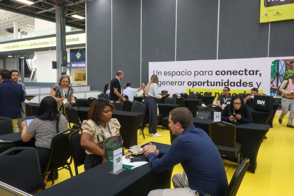 Una sala de eventos con mesas y sillas, donde varias personas interactúan y conversan. Hay un letrero en la pared que dice 'Un espacio para conectar, generar oportunidades y hacer negocios'.