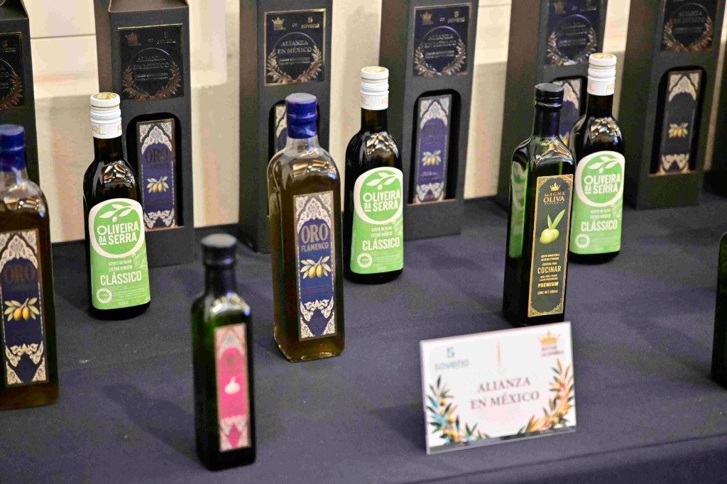 Varias botellas de aceite de oliva en una mesa, con etiquetas que indican diferentes marcas y tipos. Una tarjeta que dice 'ALIANZA EN MÉXICO' está frente a las botellas.